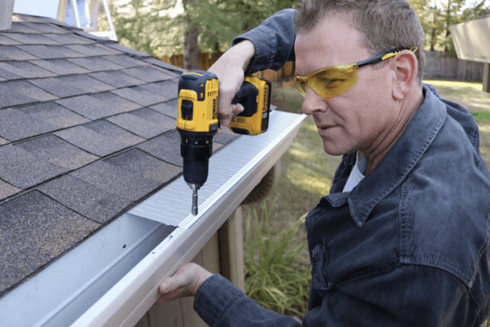 A man wearing safety glasses uses a power drill to install a Gutterglove stainless steel micro-mesh guard over a house gutter next to a shingled roof. Trees and a fence are visible in the background.